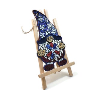 May include: A mosaic gnome ornament with a blue, red, and white design. The gnome is standing on a small wooden easel.