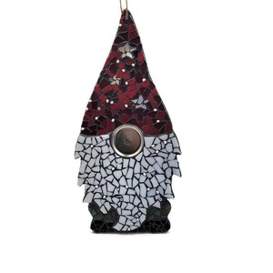 Op de afbeelding: Een moza&iuml;ek kabouter ornament met een rode en witte hoed, een zilveren knoop en een zwart-witte baard. De kabouter is gemaakt van kleine, onregelmatig gevormde stukjes glas. Er is een gouden touwtje bevestigd om op te hangen.