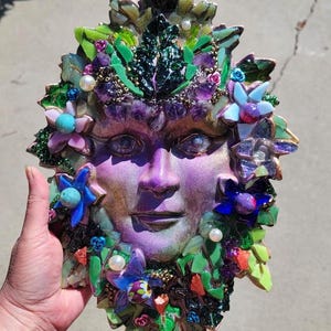 Puede incluir: Una placa decorativa de pared con un rostro morado central rodeado por un mosaico de hojas de vidrio verde, piedras de amatista moradas, detalles florales azules y rosas, y perlas blancas. El rostro presenta un acabado brillante y detallado.