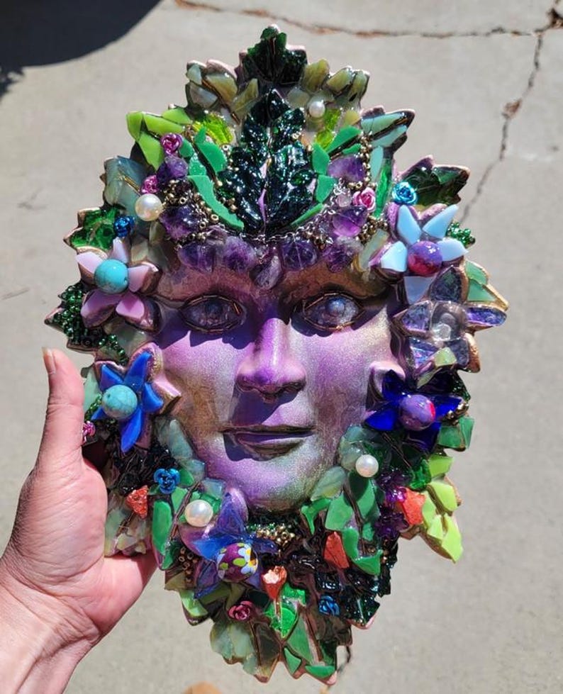 Puede incluir: Una placa decorativa de pared con un rostro morado central rodeado por un mosaico de hojas de vidrio verde, piedras de amatista moradas, detalles florales azules y rosas, y perlas blancas. El rostro presenta un acabado brillante y detallado.