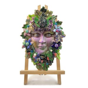 Puede incluir: Una m&aacute;scara decorativa de pared con un rostro humano central en acabado p&uacute;rpura y verde nacarado, rodeado por un mosaico de hojas verdes, peque&ntilde;as flores azules y rosas, perlas blancas y detalles de cristales de color p&uacute;rpura oscuro sobre un caballete.