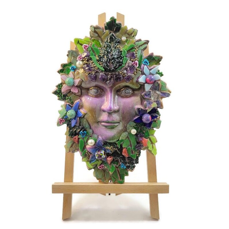 Puede incluir: Una m&aacute;scara decorativa de pared con un rostro humano central en acabado p&uacute;rpura y verde nacarado, rodeado por un mosaico de hojas verdes, peque&ntilde;as flores azules y rosas, perlas blancas y detalles de cristales de color p&uacute;rpura oscuro sobre un caballete.