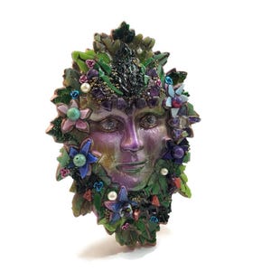 Puede incluir: Una m&aacute;scara decorativa de pared con un rostro humano central de acabado iridiscente en tonos morados y verdes. La m&aacute;scara est&aacute; rodeada por un denso mosaico de hojas verdes, peque&ntilde;as flores coloridas, perlas blancas y gemas de color morado oscuro.