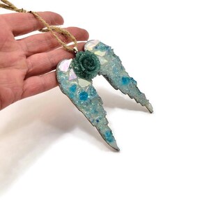 May include: A mosaic angel wing ornament with a teal rose in the center. The wings are made of iridescent glass and blue accents, attached to a natural twine cord. The ornament is a decorative item.