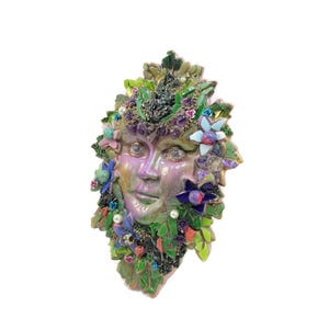 Puede incluir: Una m&aacute;scara decorativa de pared con un rostro central iridiscente en tonos morados y verdes, rodeado por un mosaico de hojas verdes, peque&ntilde;as flores coloridas, perlas blancas y detalles en morado que parecen piedras preciosas.