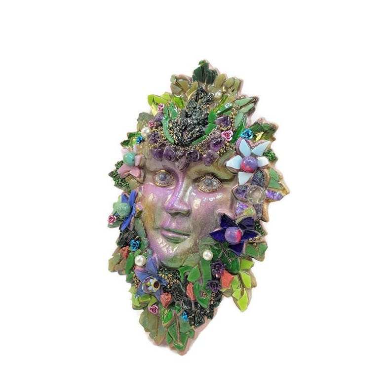 Puede incluir: Una m&aacute;scara decorativa de pared con un rostro central iridiscente en tonos morados y verdes, rodeado por un mosaico de hojas verdes, peque&ntilde;as flores coloridas, perlas blancas y detalles en morado que parecen piedras preciosas.
