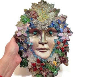 Op de afbeelding: Een wandplaat met een centraal menselijk gezichtssculptuur omringd door een mozaïek van gekleurde glasscherven, kleine bloemvormen en een zilverkleurig vlinderornament in het midden boven het voorhoofd.
