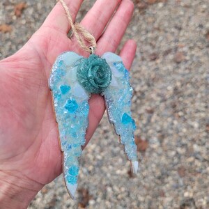May include: A handcrafted angel wing ornament with a teal rose center. The wings are made of wood and decorated with blue and white glass pieces. A twine loop is attached for hanging. The ornament is a unique, decorative item.