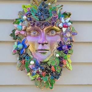 Puede incluir: Una escultura de m&aacute;scara para pared con un rostro iridiscente en tonos morados y dorados, rodeado por un mosaico de hojas de vidrio verde, piedras de amatista moradas, perlas blancas y varios acentos florales coloridos en azul, rosa y blanco.