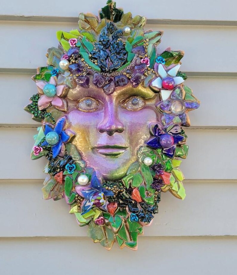 Puede incluir: Una escultura de m&aacute;scara para pared con un rostro iridiscente en tonos morados y dorados, rodeado por un mosaico de hojas de vidrio verde, piedras de amatista moradas, perlas blancas y varios acentos florales coloridos en azul, rosa y blanco.