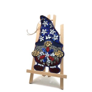 May include: Mosaic gnome ornament with a red, white, and blue hat and beard. The gnome is standing on a wooden easel.