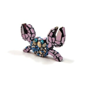 May include: A pink mosaic crab sculpture with blue and white accents. The crab has large, round eyes and is decorated with small shells and beads.