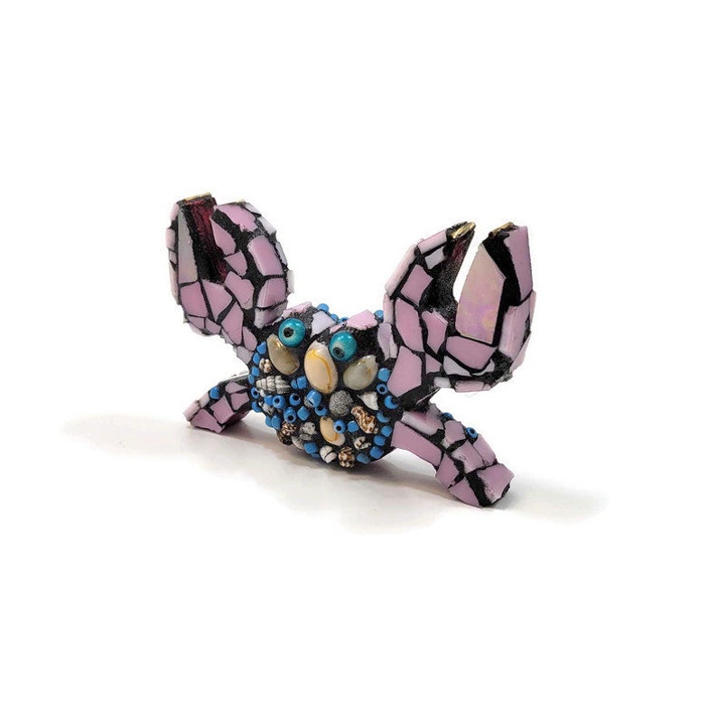 May include: A pink mosaic crab sculpture with blue and white accents. The crab has large, round eyes and is decorated with small shells and beads.