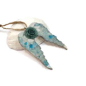 May include: A handcrafted angel wing pendant with a teal rose center. The wings are light blue with iridescent accents and embedded blue glass pieces. The pendant is attached to a natural twine cord, perfect for wearing or displaying.