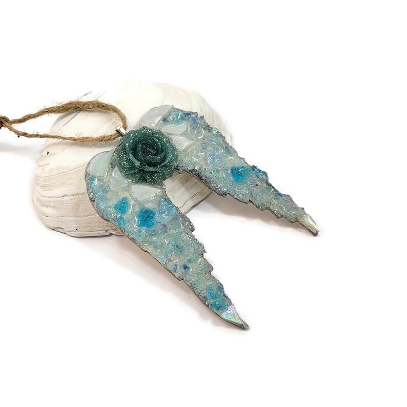 May include: A handcrafted angel wing pendant with a teal rose center. The wings are light blue with iridescent accents and embedded blue glass pieces. The pendant is attached to a natural twine cord, perfect for wearing or displaying.