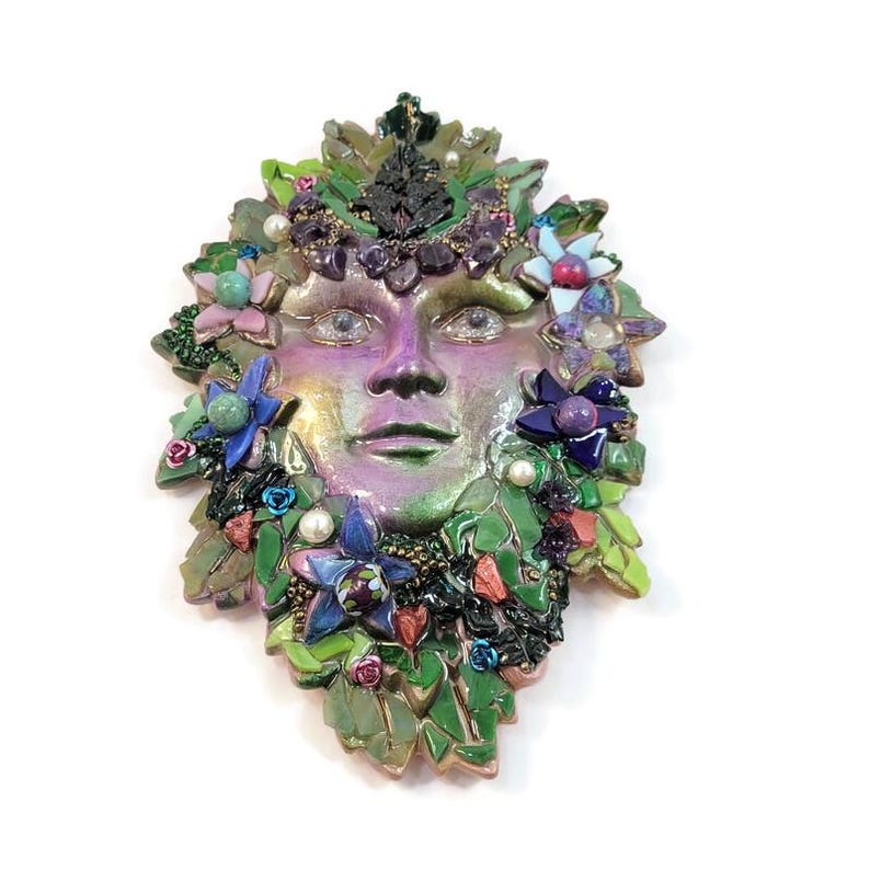 Puede incluir: Una m&aacute;scara decorativa de pared con un rostro humano central en tonos p&uacute;rpuras y verdes iridiscentes. El rostro est&aacute; rodeado por un mosaico de hojas verdes, peque&ntilde;as flores azules y rosas, perlas blancas y detalles de piedras moradas oscuras.