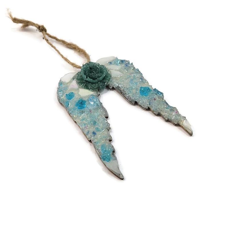 May include: A decorative angel wing ornament with a teal rose in the center. The wings are light blue with white and teal accents, and are attached to a natural twine string. The ornament is set against a white background.