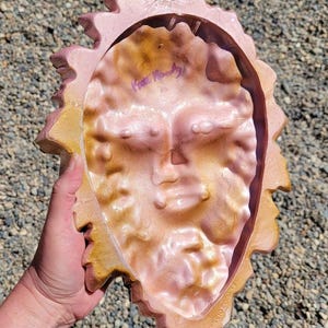 Puede incluir: Una placa decorativa de pared con forma de cara de sol y un borde festoneado similar a una hoja. La pieza presenta un degradado de tonos oro met&aacute;lico y rosa suave. Una firma est&aacute; escrita con tinta morada en la zona de la frente del sol.