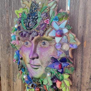 Puede incluir: Una escultura de m&aacute;scara de pared que presenta un rostro humano en tonos morados y verdes, adornada con un mosaico de fragmentos de vidrio de colores, peque&ntilde;as cuentas, piedras iridiscentes y detalles en forma de flor en azul, rosa y blanco.