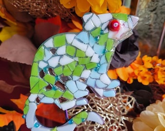 Op de afbeelding: Een mozaïek tuinpen in de vorm van een muis, gemaakt van kleine, onregelmatige stukjes wit, lichtblauw en limoengroen glas. De muis heeft een opvallende rode glazen kraal als oog en is versierd met kleine blauwe kraaltjes.