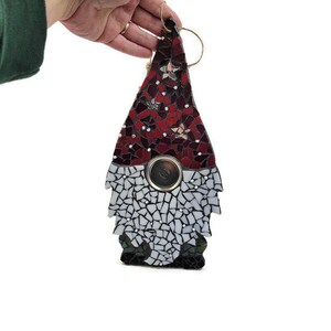 Op de afbeelding: Een moza&iuml;ek kabouter ornament in rood en wit. De hoed van de kabouter is gemaakt van rode moza&iuml;ektegels, terwijl de baard wit is. Een metalen cirkel bevindt zich in het midden van het lichaam van de kabouter. Een touwlus is aan de bovenkant bevestigd om op te hangen.