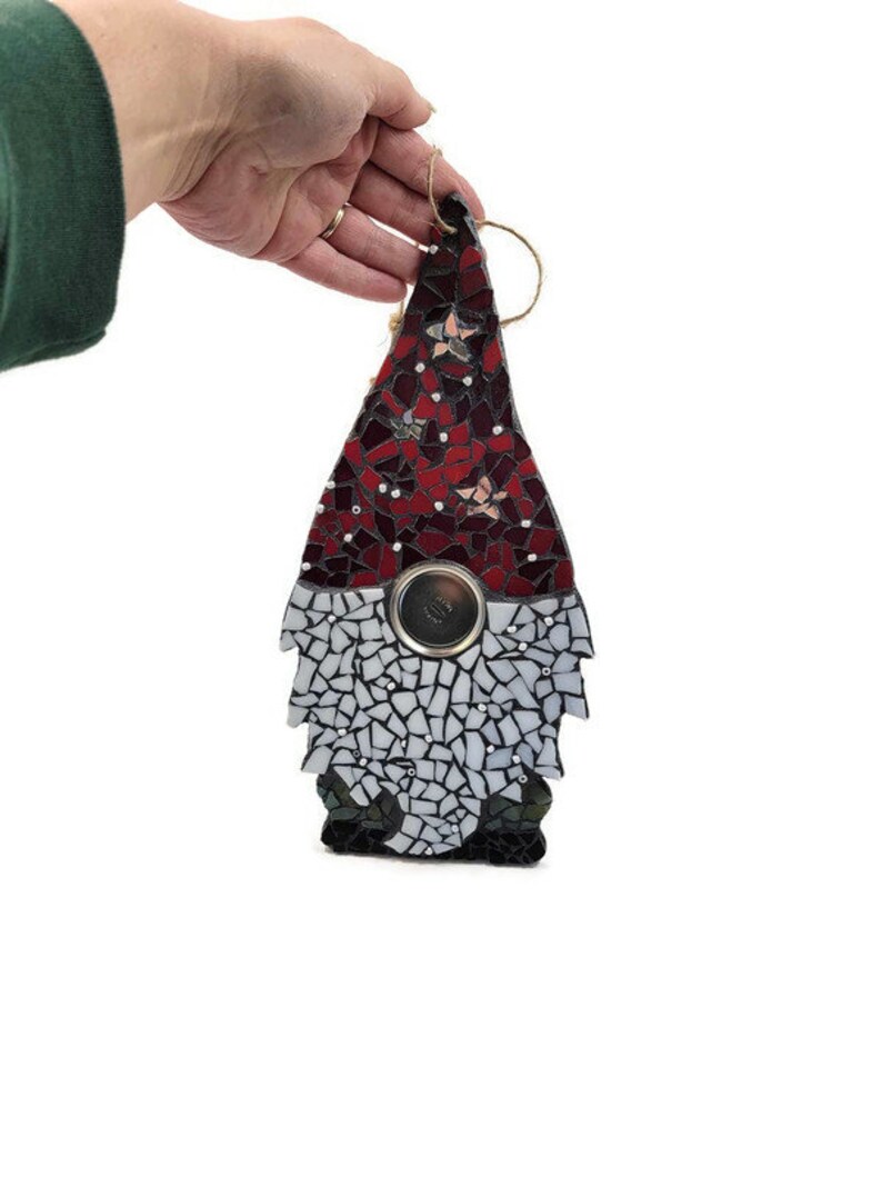 Op de afbeelding: Een moza&iuml;ek kabouter ornament in rood en wit. De hoed van de kabouter is gemaakt van rode moza&iuml;ektegels, terwijl de baard wit is. Een metalen cirkel bevindt zich in het midden van het lichaam van de kabouter. Een touwlus is aan de bovenkant bevestigd om op te hangen.