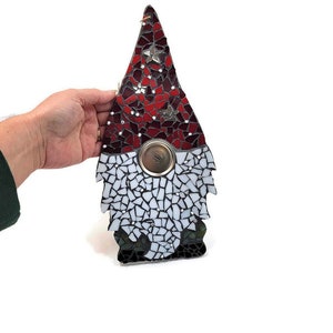 Op de afbeelding: Een moza&iuml;ek kabouterbeeldje met een rode en witte puntmuts, een witte baard en een cirkelvormig metalen element. De hoed heeft stervormige accenten. De baard van de kabouter is gemaakt van witte moza&iuml;ektegels.