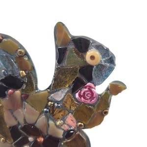 Puede incluir: Una escultura de mosaico de ardilla con una rosa rosa y varios trozos de vidrio de colores. La ardilla tiene un ojo marrón claro y sostiene una rosa. El mosaico está hecho de varios colores, incluyendo marrón, verde y azul.