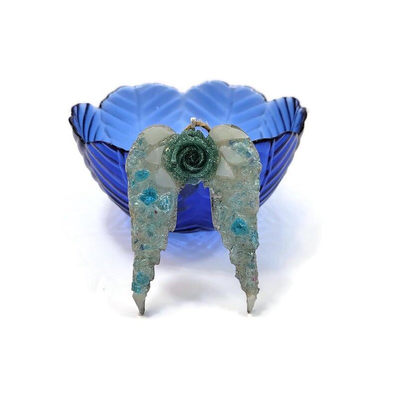 May include: A handcrafted angel wing pendant with a green rose accent. The wings are made of clear resin with blue and white accents. The pendant is displayed in front of a blue glass bowl.