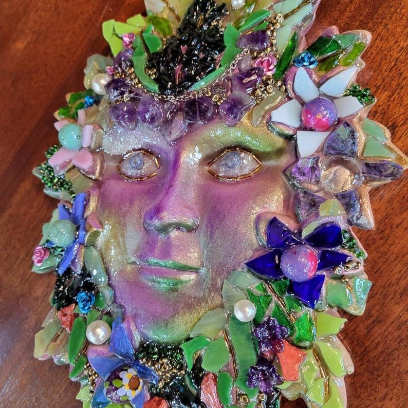 Puede incluir: Una m&aacute;scara decorativa de pared con un rostro humano en acabado iridiscente morado y verde. La m&aacute;scara est&aacute; adornada con un mosaico de fragmentos de vidrio de colores, piedras moradas tipo amatista, perlas blancas y detalles de flores de vidrio.