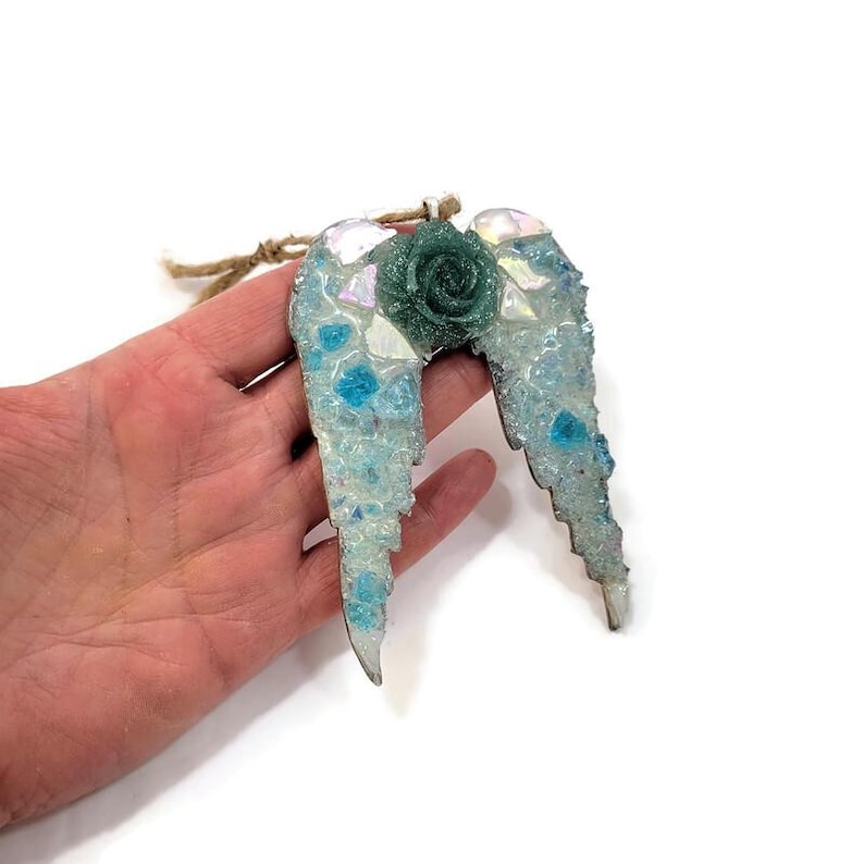 May include: A decorative angel wing ornament with a teal rose in the center. The wings are light blue with iridescent and blue accents. A twine loop is attached for hanging. The ornament is approximately 4 inches wide.
