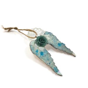 May include: A decorative ornament shaped like angel wings. The wings are light blue with a mosaic of clear and blue glass pieces. A green rose is at the center, and a twine loop is attached for hanging.