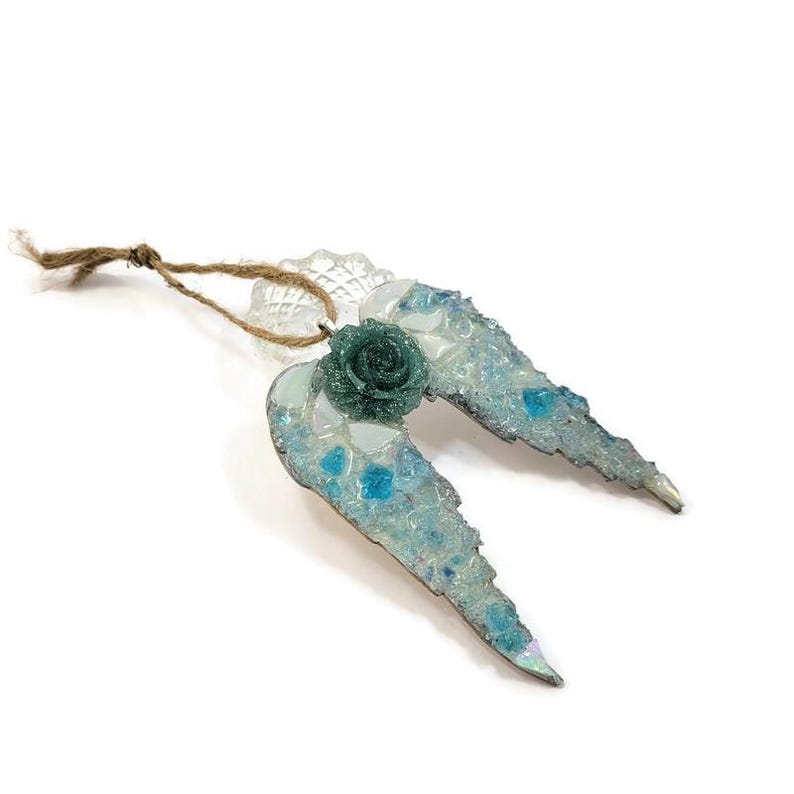 May include: A decorative ornament shaped like angel wings. The wings are light blue with a mosaic of clear and blue glass pieces. A green rose is at the center, and a twine loop is attached for hanging.