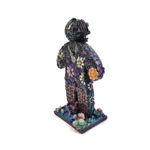 May include: A mosaic figurine of a person with a black head and purple, orange, and white flowers on their torso. The figurine is standing on a mosaic base with blue, green, and purple accents.
