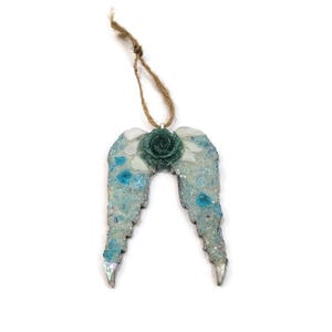 May include: A decorative angel wing ornament with a teal rose center and a jute string for hanging. The wings are a mosaic of light blue, white, and clear glass pieces. The ornament is approximately 4 inches wide.