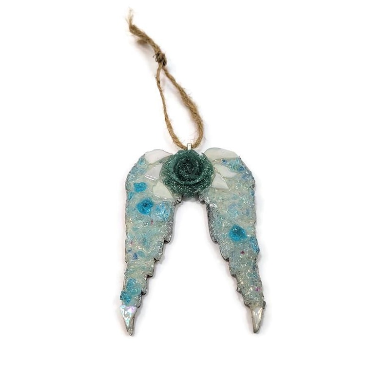 May include: A decorative angel wing ornament with a teal rose center and a jute string for hanging. The wings are a mosaic of light blue, white, and clear glass pieces. The ornament is approximately 4 inches wide.