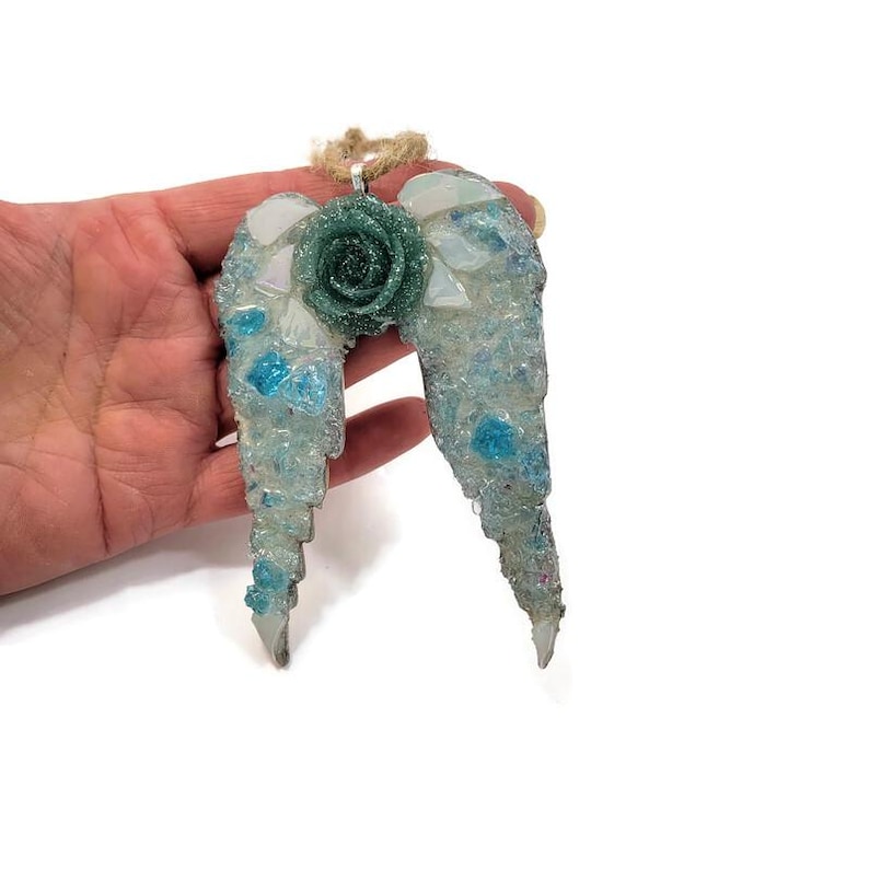 May include: A decorative angel wing ornament with a teal rose in the center. The wings are made of clear and blue glass pieces, with a silver-colored metal loop and a natural twine hanger.