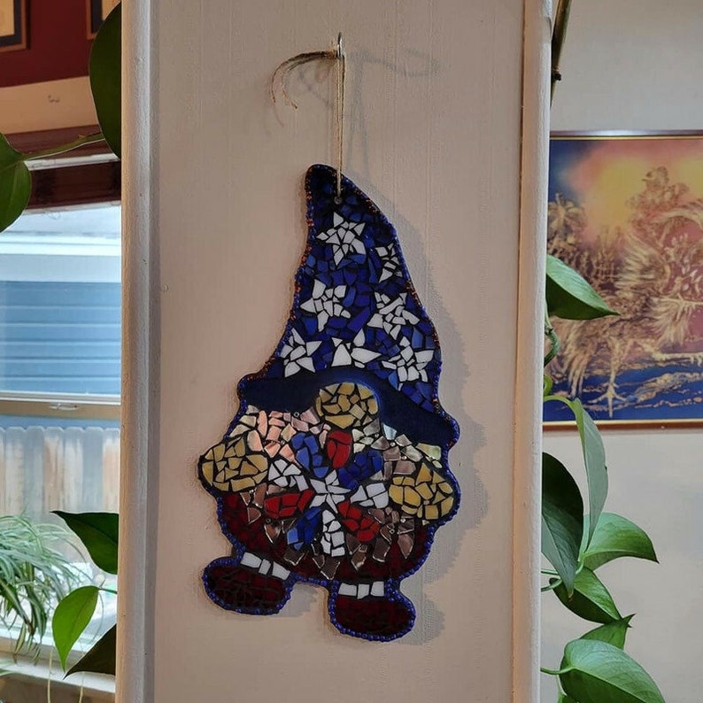 May include: A mosaic gnome ornament with a blue, white, and red hat and a red, white, and blue body. The gnome is standing on a white surface.