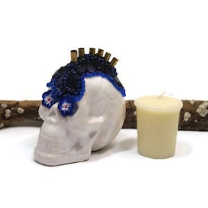 May include: White ceramic skull with blue and black beaded embellishments on the top of the head. The skull is sitting on a piece of driftwood with a small white candle next to it.