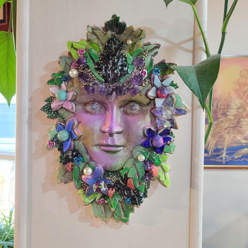 Puede incluir: Una escultura de pared en forma de m&aacute;scara con un rostro humano central de acabado iridiscente p&uacute;rpura y verde. La m&aacute;scara est&aacute; rodeada por un mosaico de hojas de vidrio verde, piedras de amatista moradas, peque&ntilde;as perlas blancas y flores.