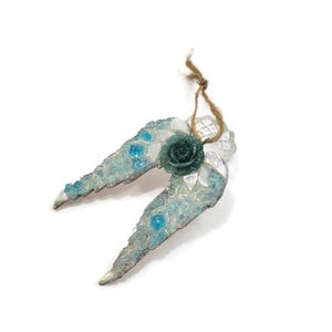May include: A decorative angel wing ornament with a teal glitter rose in the center. The wings are light blue with clear and teal accents, and a jute string for hanging. The ornament is approximately 4 inches wide.