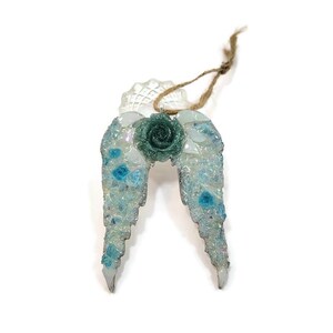 May include: A decorative angel wing ornament with a teal rose in the center. The wings are made of clear and blue mosaic pieces, with a clear, scalloped top and a jute string for hanging.