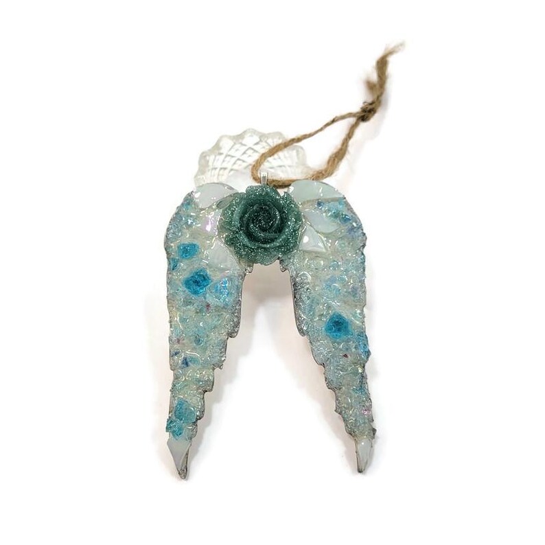 May include: A decorative angel wing ornament with a teal rose in the center. The wings are made of clear and blue mosaic pieces, with a clear, scalloped top and a jute string for hanging.