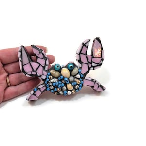 May include: A pink mosaic crab with blue and white eyes, decorated with seashells and beads.