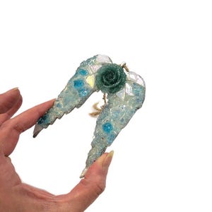 May include: A pair of decorative angel wings with a teal rose in the center. The wings are light blue with iridescent and blue accents. The wings are held by a hand against a white background.
