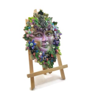 Puede incluir: Una m&aacute;scara decorativa de pared con un rostro humano de acabado iridiscente en morado y verde, rodeado por un mosaico de hojas verdes, flores azules y rosas, peque&ntilde;as perlas blancas y piedras de color morado oscuro, sobre un caballete de madera.