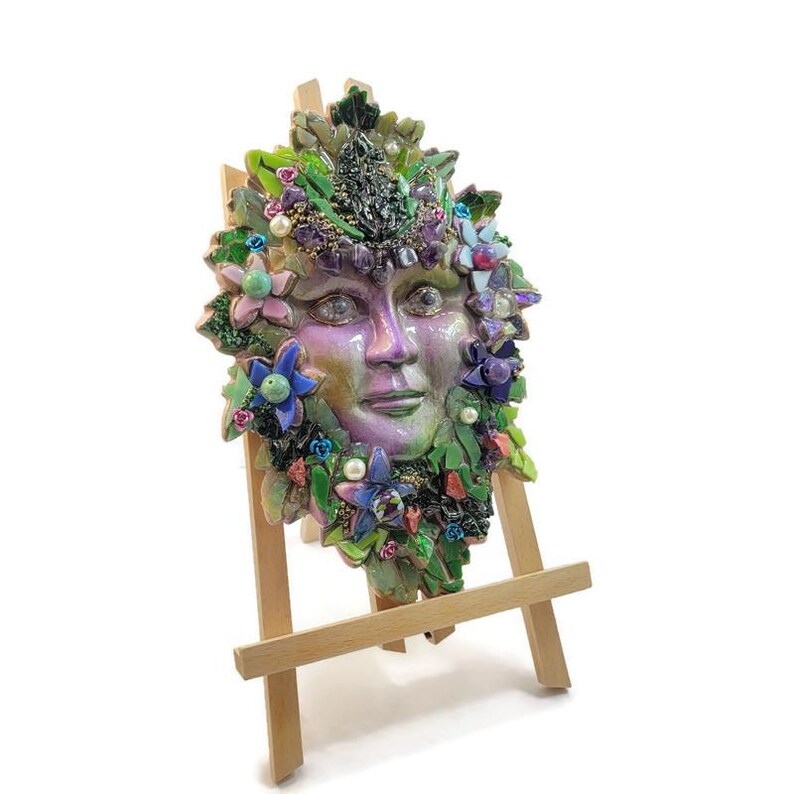 Puede incluir: Una m&aacute;scara decorativa de pared con un rostro humano de acabado iridiscente en morado y verde, rodeado por un mosaico de hojas verdes, flores azules y rosas, peque&ntilde;as perlas blancas y piedras de color morado oscuro, sobre un caballete de madera.