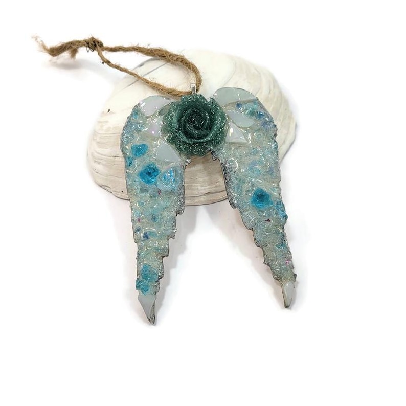 May include: A handcrafted angel wing ornament with a teal rose center. The wings are light blue with embedded blue and clear accents. A natural twine loop is attached for hanging. The ornament is resting on a white seashell.