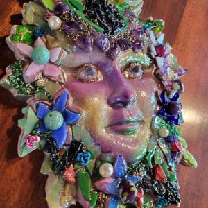 Puede incluir: Una m&aacute;scara decorativa de pared con un rostro humano central en acabado p&uacute;rpura y dorado nacarado. La m&aacute;scara est&aacute; adornada con un mosaico de fragmentos de vidrio de colores, peque&ntilde;as hojas verdes, amatistas, detalles florales azules y perlas blancas.