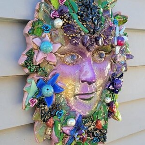 Puede incluir: Una m&aacute;scara decorativa de pared con un rostro central de color p&uacute;rpura iridiscente rodeado por un mosaico de fragmentos de vidrio verde, azul y rosa, peque&ntilde;as flores artificiales y cuentas de perlas blancas. La pieza tiene un aspecto texturizado.